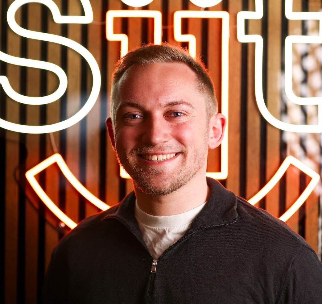 Man smiling in front of neon sign.
